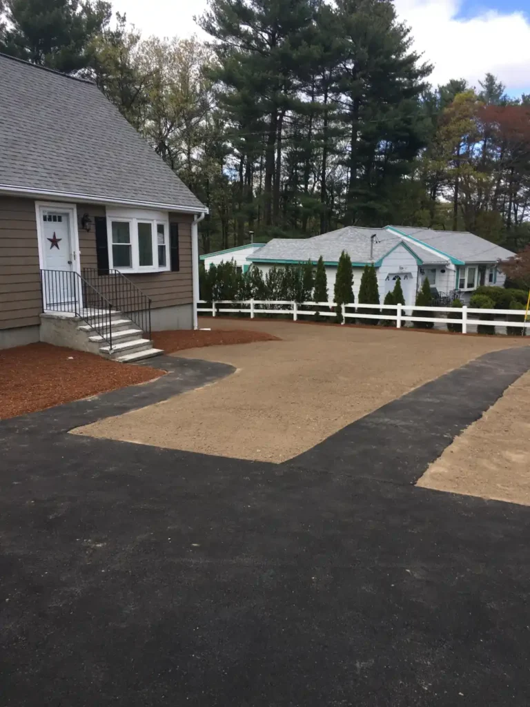 Residential landscaping project showcasing a newly prepared walkway area with compacted gravel, adjacent to a home in Massachusetts, surrounded by trees and neighboring properties.