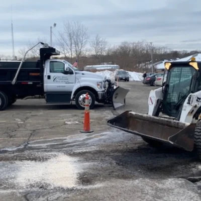 Commercial snow management equipment including a Ford truck with a plow and a Bobcat loader, positioned on a snow-covered parking lot, demonstrating effective snow removal and salt application services.