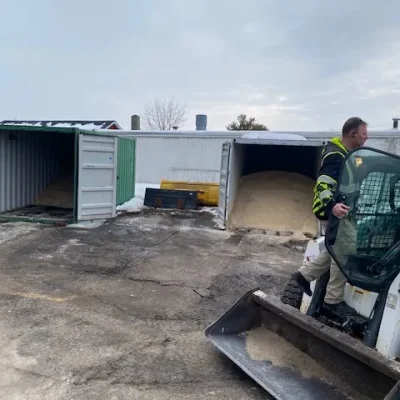 Bobcat operator preparing for snow management, with salt storage containers in the background, illustrating commercial snow management services.